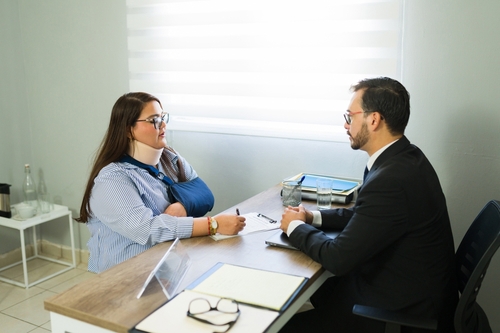 Woman with arm sling meeting attorney across desk with clipboard and documents