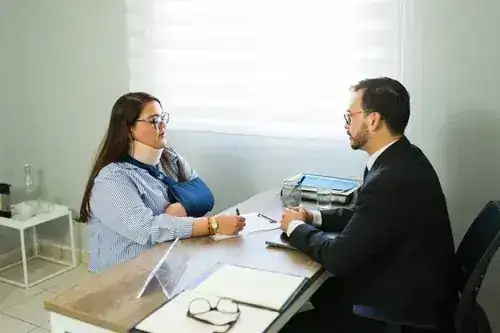 Woman with arm sling meeting attorney across desk with clipboard and documents